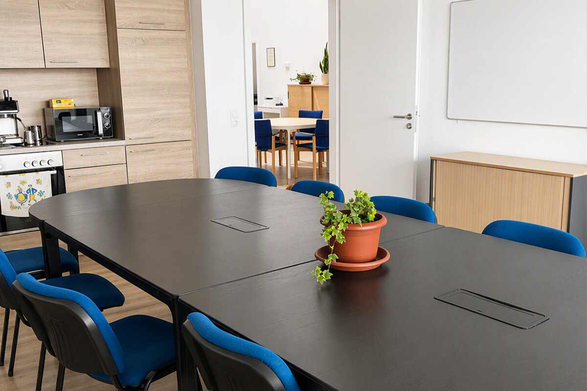 Schwarzer Konferenztisch mit blauen Stühlen und einer Pflanze in der Mitte in einem hellen Raum mit Küchenzeile im Hintergrund