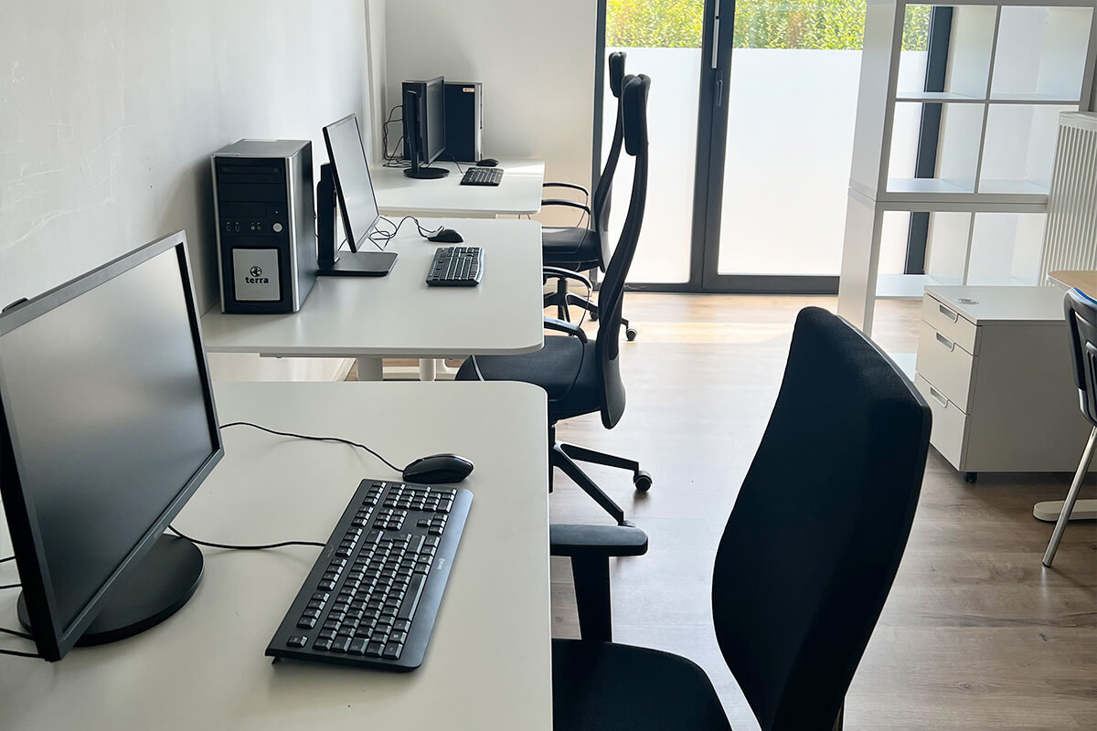 Raum mit mehreren weißen Schreibtischen, schwarzen Bürostühlen, Computern und einer großen Fensterfront im Hintergrund